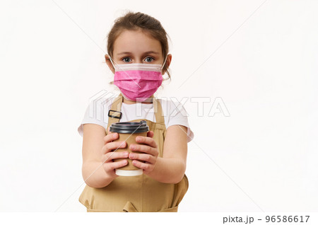 Caucasian little child girl wearing safety medical mask, dressed as barista, bartender with a hot drink in paper cup, isolated on white background. Cafe restaurant on pandemic lockdown. Takeaway food. Caucasian little child girl wearing safety medical mask, dressed as barista, bartender with a hot drink in paper cup, isolated on white background. Cafe restaurant on pandemic lockdown. Takeaway food. 96586617