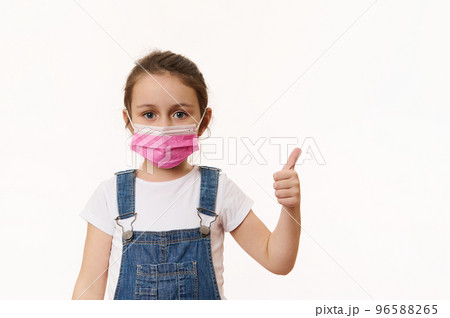 Cute child girl in blue denim overalls, wearing a pink safety mask, shows thumb up, looking at camera, isolated over white background. Space for promo text. Pediatrics. Health care and medical content Cute child girl in blue denim overalls, wearing a pink safety mask, shows thumb up, looking at camera, isolated over white background. Space for promo text. Pediatrics. Health care and medical content 96588265