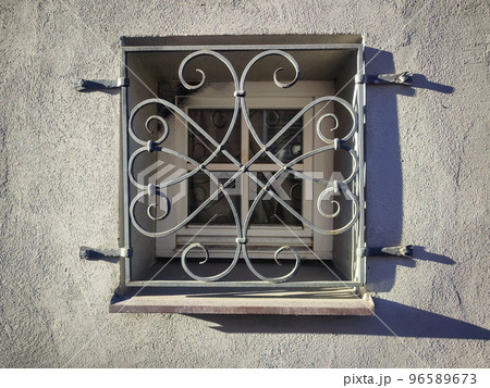 Beautiful old square window forged window security grate with long shadows on concrete building wall on sunny evening 96589673