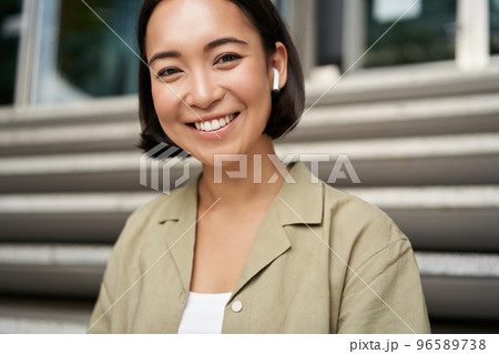 Portrait of smiling asian girl listens music, podast in wireless earphones, using headphones outdoors, sitting on street 96589738