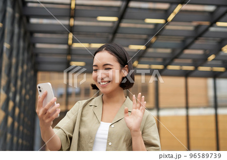 Happy asian girl waves at her smartphone camera, says hello to friend on video chat, calling someone, standing on street 96589739