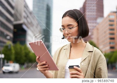 Portrait of happy young woman in glasses, standing on street with cup of coffee and tablet Portrait of happy young woman in glasses, standing on street with cup of coffee and tablet 96589740
