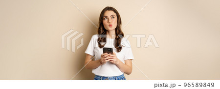 Portrait of woman thinking, holding mobile phone and looking up with thoughtful face, standing over beige background 96589849