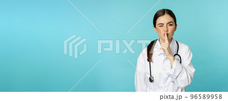 Smiling cute doctor woman, shush, press hush shh sign with finger pressed to lips, hiding secret, taboo gesture, standing over torquoise background Smiling cute doctor woman, shush, press hush shh sign with finger pressed to lips, hiding secret, taboo gesture, standing over torquoise background 96589958