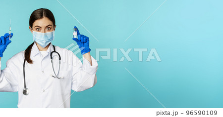 Smiling medical staff, doctor in face mask and rubber gloves, showing syringe and vaccine from covid-19 pandemic, standing over blue background 96590109