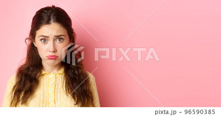 Close up of cute and coy girl making sad pouting face to ask for something, wants pity, standing against pink background gloomy Close up of cute and coy girl making sad pouting face to ask for something, wants pity, standing against pink background gloomy 96590385