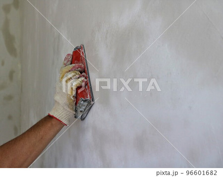 sanding the surface of a plastered wall with a hand sander close-up, a plasterer's hand brushing off the unevenness of the wall after applying the mortar and copy space for text 96601682