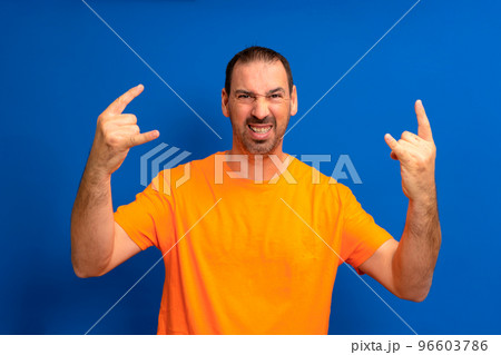 Bearded latino man wearing orange t-shirt with crazy expression doing rock symbol with hands up. Music star heavy concept. 96603786