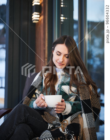 Young woman resting on terrace of modern barn house in the mountains. Happy female tourist sitting in chair, holding cup of tea, enjoying in new cottage in winter. 96604332