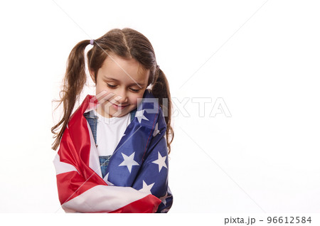 Close-up of delightful little girl with two cute ponytails, hugs herself, covered on United States of America flag, on independence day, isolated over white background, with space for your advertising 96612584