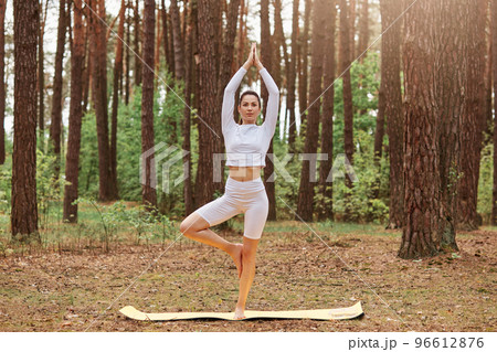 Outdoor full length portrait of sporty woman standing on one leg and stretching arms up, pressing palms together, female in tree yoga pose, practicing in forest. 96612876