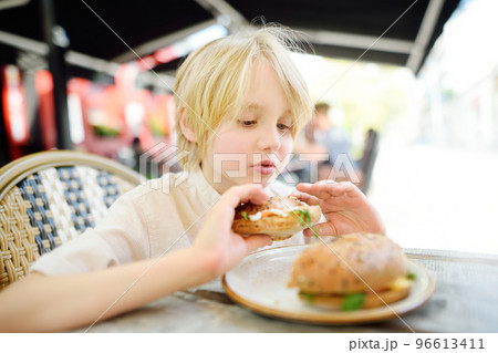 Cute blonde boy eating large bagel with salmon and arugula in outdoor fast food restaurant 96613411