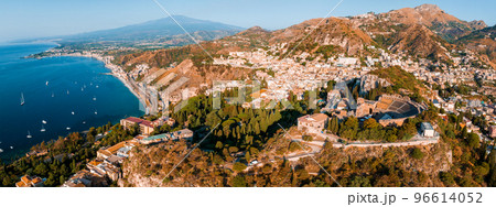Aerial view of the Ruins of the Ancient Greek Theater in Taormina, Sicily with the double smoke tail of the Etna extending over the the Giardini-Naxos bay of the Ionian Sea in the morning sun shine. Aerial view of the Ruins of the Ancient Greek Theater in Taormina, Sicily with the double smoke tail of the Etna extending over the the Giardini-Naxos bay of the Ionian Sea in the morning sun shine. 96614052