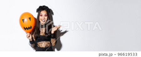 Image of beautiful asian woman celebrating halloween, wearing witch costume and gothic makeup, talking with orange balloon Image of beautiful asian woman celebrating halloween, wearing witch costume and gothic makeup, talking with orange balloon 96619533