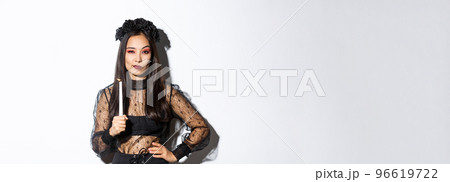 Young sassy witch practice magic with lit candle on halloween, wearing gothic lace dress and looking at camera confident, standing over white background Young sassy witch practice magic with lit candle on halloween, wearing gothic lace dress and looking at camera confident, standing over white background 96619722