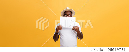 Young happy African-american hiding behind a blank paper, isolated on yellow background 96619781