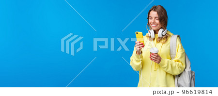 Girl using mobile phone to contact friend after college, texting as walking along street with backpack, take-away coffee cup and headphones, smiling joyfully smartphone display Girl using mobile phone to contact friend after college, texting as walking along street with backpack, take-away coffee cup and headphones, smiling joyfully smartphone display 96619814
