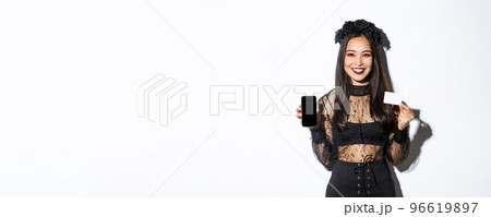Smiling attractive woman in black gothic dress showing credit card and smartphone display, standing over white background in halloween costume Smiling attractive woman in black gothic dress showing credit card and smartphone display, standing over white background in halloween costume 96619897