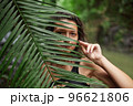 Portrait Young slender girl posing in the jungle hiding behind a palm leaf. Wildlife and a girl 96621806