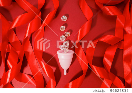 Pink menstrual cup, transparent crystals like blood drops and roll red ribbons on maroon red background. Women's health, alternative hygiene and ecology of planet. Top front view, copy space, flat lay 96622355