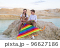 A boy and a girl are having fun sitting near the river holding a bright striped kite 96627186