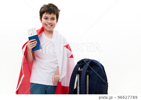 Multiethnic happy smart teenage boy in casual t-shirt and blue jeans, smiling, looking at camera, posing with Canadian flag, boarding pass and backpack on white background. Free space for advertising 96627789