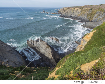 View of sea shore with small wooden bridge, ocean waves, cliffs and sand dunes covered by green vegetation red leaves of sour fig, sunny day, clear blue sky. Rota Vicentina coast, Portugal 96628135