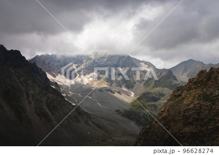 Dramatic view of forested high rocky slopes and snowy mountains in gray cloudy sky in changeable weather, mountain landscape without people 96628274