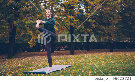 Female yoga student is balancing on one leg standing on mat in recreational area exercising alone on warm autumn day. Modern youth, healthcare and nature concept. 96628721