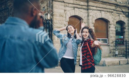 African American man tourist is taking pictures of his pretty female friends posing for camera. Tourism, photography and youth concept. African American man tourist is taking pictures of his pretty female friends posing for camera. Tourism, photography and youth concept. 96628830