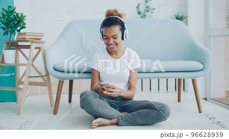 Pretty girl is playing game on smartphone touching screen and smiling while listening to music with headphones sitting on floor at home. Technology, millennials and fun concept. 96628939