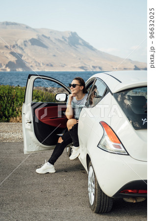 A beautiful brunette in sunglasses sits by a white car against the backdrop of the Mediterranean Sea and mountains A beautiful brunette in sunglasses sits by a white car against the backdrop of the Mediterranean Sea and mountains 96632025