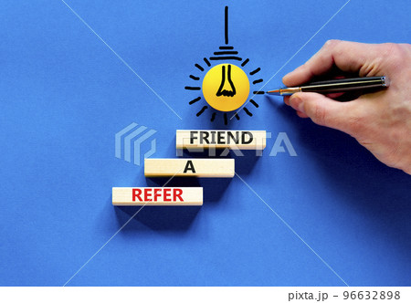 Refer a friend symbol. Concept words Refer a friend on wooden blocks on a beautiful blue table blue background. Yellow light bulb. Businessman hand. Business and refer a friend concept. Copy space. 96632898