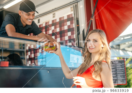 Cook in a food truck handing tasty burger over to woman customer Cook in a food truck handing tasty burger over to woman customer 96633919