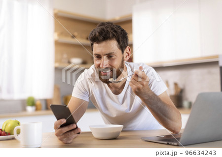 Smiling mature caucasian male with beard in white t-shirt eats porridge, read news on smartphone, watches video Smiling mature caucasian male with beard in white t-shirt eats porridge, read news on smartphone, watches video 96634243