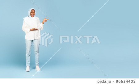 Happy black woman in hijab pointing aside at free space for your ad, standing over blue background, panorama Happy black woman in hijab pointing aside at free space for your ad, standing over blue background, panorama 96634405