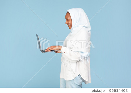 African american muslim woman typing on computer keyboard and smiling over blue studio background, side view, copy space 96634408