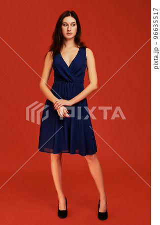 Beautiful young brunette woman in elegant blue cocktail dress and black high heels is posing for the camera Beautiful young brunette woman in elegant blue cocktail dress and black high heels is posing for the camera 96635517