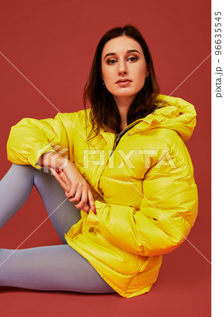 Studio portrait of young brunette woman sitting on the floor in yellow down jacket and grey blue panty hoses or stockings Studio portrait of young brunette woman sitting on the floor in yellow down jacket and grey blue panty hoses or stockings 96635545