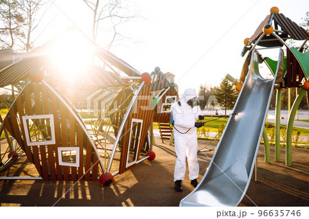 Man wearing protective suit disinfecting the playground in the sun with spray. Covid -19. Cleaning. 96635746