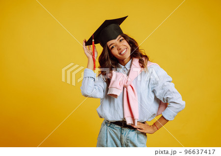 Graduate woman in a graduation hat on her head posing on a yellow background. Study, education, university, college, graduate concept. 96637417