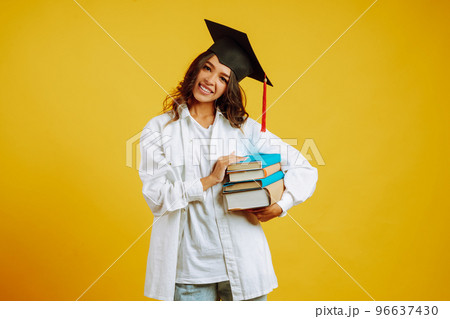 Graduate woman in a graduation hat on her head, with books stands on a yellow background. 96637430