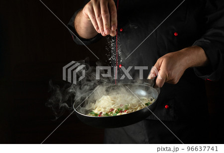 The chef adds salt to the spaghetti in a steaming hot pan. Recipe or menu for restaurant or hotel on black background 96637741