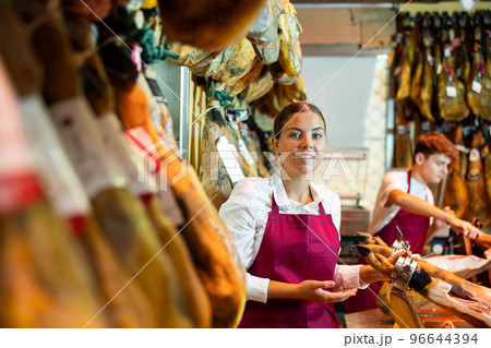 Young seller arranging jamon at counter in butcher shop Young seller arranging jamon at counter in butcher shop 96644394