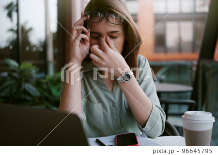 A tired and stressed young woman holds glasses and massages his nose while working hard in a cafe. A tired and stressed young woman holds glasses and massages his nose while working hard in a cafe. 96645795