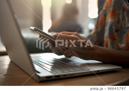 Close-up of woman's hand using smartphone while working on laptop in a coffee shop. 96645797