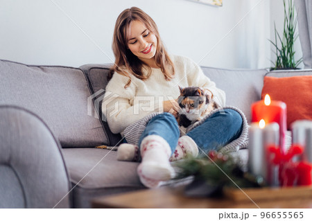 Woman in plaid with tea cup watching movie, TV, petting cat on sofa at home with christmas decoration atmosphere. Lady wear jumper and warm socks. Cozy and comfortable winter concept. Selective focus. 96655565