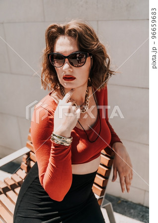 Portrait of a woman on the street. An attractive woman in glasses, a red blouse and a black skirt is sitting on a bench outside. Portrait of a woman on the street. An attractive woman in glasses, a red blouse and a black skirt is sitting on a bench outside. 96659683