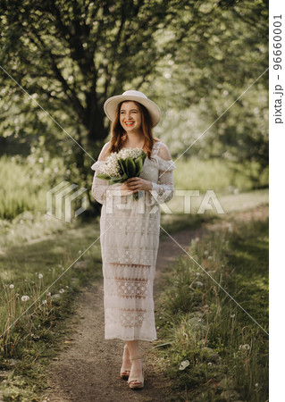 Portrait of a beautiful woman in a white dress and a hat with lilies of the valley. A girl in nature. Spring flowers 96660001