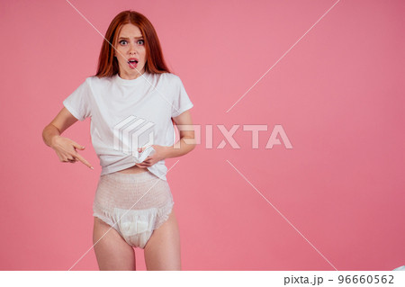 young amazed and surprised redhaired ginger woman wearing incontinence diaper in studio pink background.feeling shame and embarrassment 96660562